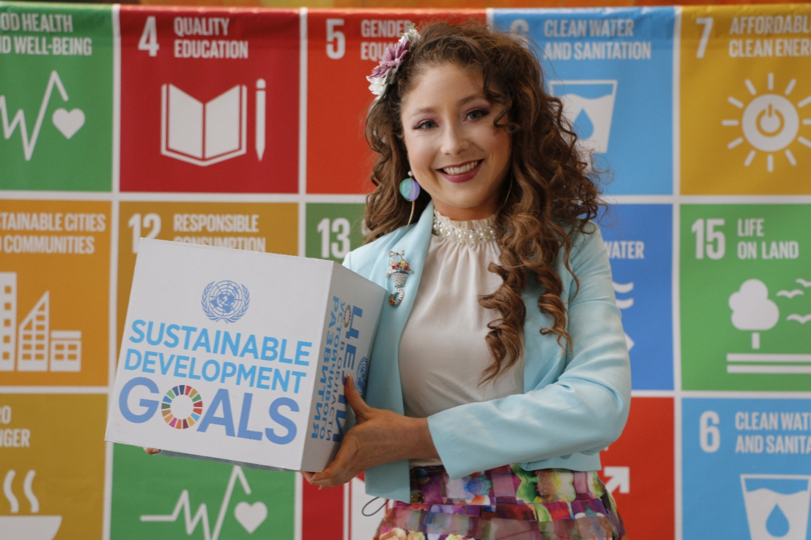 Kelly Lovell standing on stage at the United Nations with the colourful SDGs behind her as a backdrop